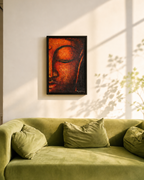 A framed wall art print featuring a serene Buddha face in warm orange and brown tones, displayed above a green velvet sofa in a sunlit room with leaf shadows on the wall.