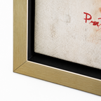 Close-up of a framed art print showing the corner of a light wood frame, black matting, textured off-white fabric background, and a partial red painted letter on a white studio background.