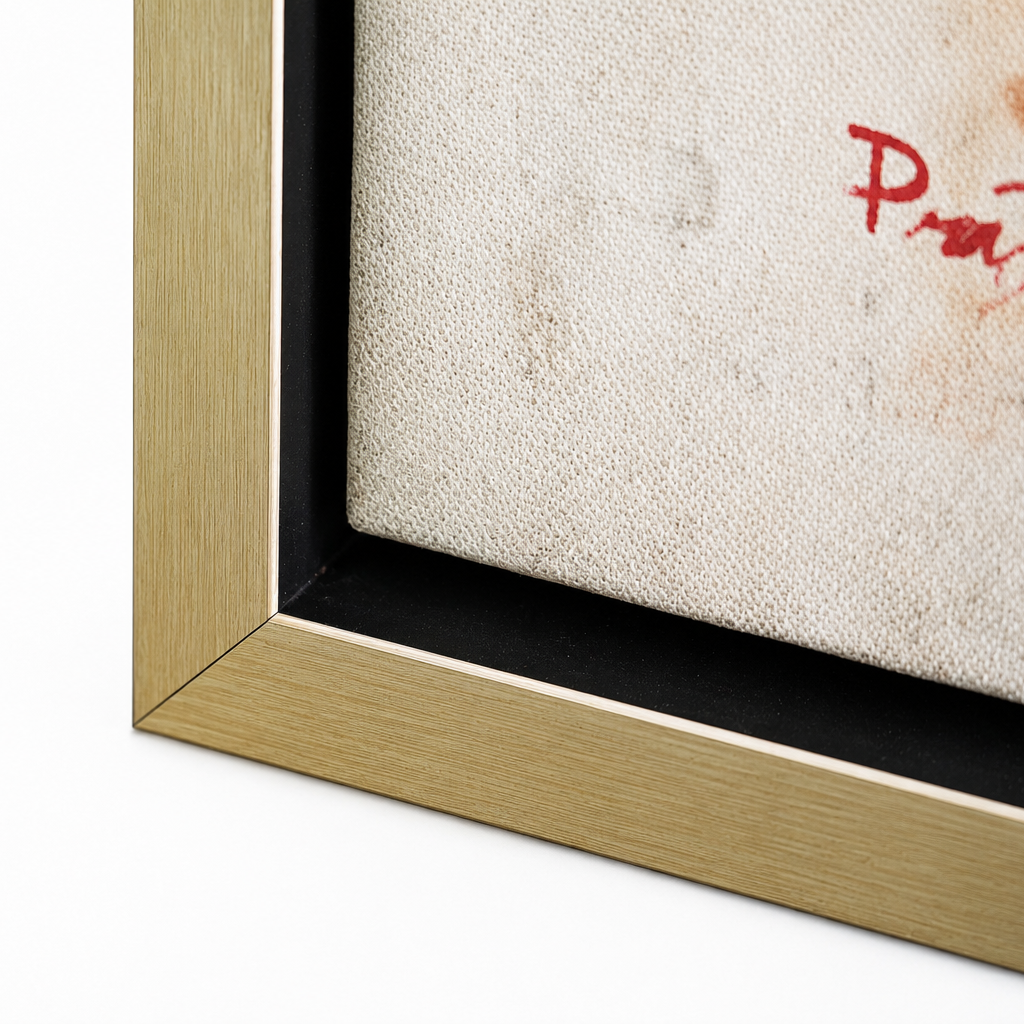 Close-up of a framed art print showing the corner of a light wood frame, black matting, textured off-white fabric background, and a partial red painted letter on a white studio background.